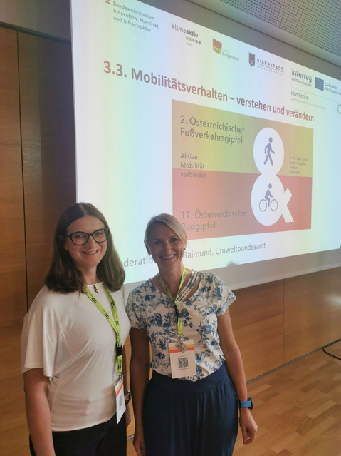 Sandra Wegener (links) mit Brille im weißen T-Shirt und Juliane Stark (rechts) im gemusterten Kurzarmshirt stehen lächelnd mit Konferenz-Lanyards vor einer Projektionswand mit der Präsentationsfolie des Rad- und Fußverkehrsgipfels.   