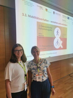Sandra Wegener (links) mit Brille im weißen T-Shirt und Juliane Stark (rechts) im gemusterten Kurzarmshirt stehen lächelnd mit Konferenz-Lanyards vor einer Projektionswand mit der Präsentationsfolie des Rad- und Fußverkehrsgipfels.   