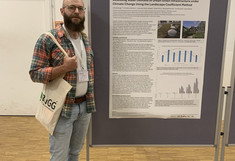 Udo Buttinger stands next to his poster, wearing a checkered shirt and jeans.