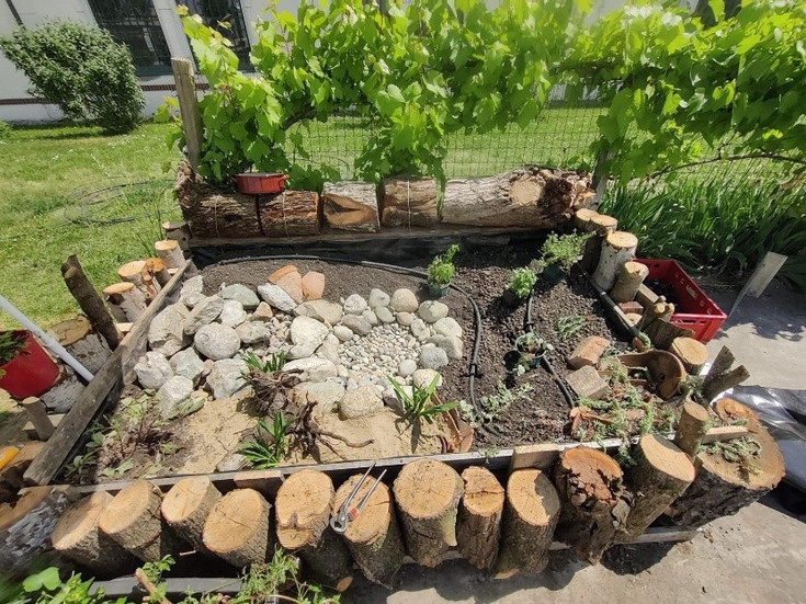 Mit senkrechten ca 60 cm honen Baumstämmen eingefasstes  Beet. Mit Wasserfläche, Steinen und Pflanzen