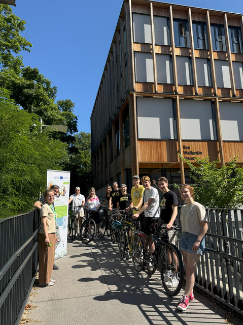 In the photo, 11 people are standing in front of a wooden building (Ilse-Wallentin-Haus). Eight of them are with bicycles. A poster of the Greenpath project is also visible.