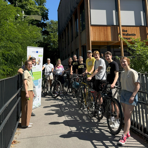 Auf dem Foto sind 11 Personen vor einem Holzhaus (Ilse-Wallentin-Haus) zu sehen. Acht von ihnen stehen mit Fahrrädern. Außerdem ist ein Poster des Projekts Greenpath im Bild zu erkennen.