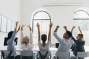 Woman in formal shirt with blonde hair waving hands, sitting between coworkers in big light conference hall. Photo from back of tired managers stretching during meeting in office..