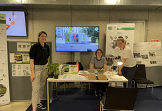 Drei Frauen am Infostand, seitlich Plakate, dahinter ein Info-Monitor.