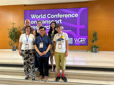Sechs Forschende des Instituts für Verkehrswesen lächeln gemeinsam auf einer Konferenzbühne vor einem großen violetten Bildschirm mit der Aufschrift „World Conference on Transport Research“. In der hinteren Reihe stehen von links nach rechts Juliane Stark, Georgia Charalampidou und Liting Yuan. In der vorderen Reihe stehen von links nach rechts Astrid G., Yusak Susilo und Paul Pfaffenbichler.