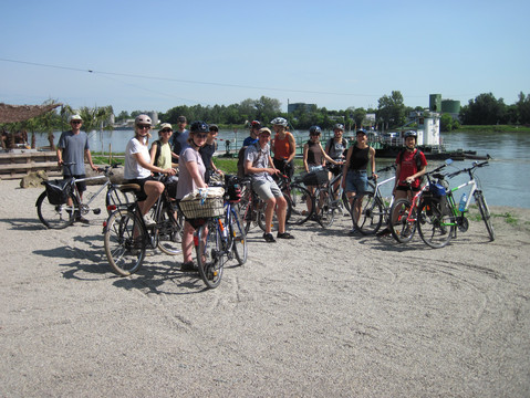 Auf dem Foto sind Studenten-Teilnehmer mit ihren Fahrrädern an einem Fluss. Sie machen eine Pause und schauen in die Kamera.