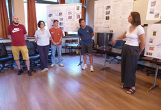 Eine Studentin steht rechts im Bild vor zwei großen Postern und erklärt gestikulierend die Ergebnisse. Links im Hintergrund stehen vier weitere Teammitglieder an der Wand und verfolgen aufmerksam ihre Ausführungen.
