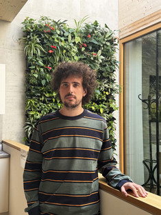 Porträt von Lorenzo Maccarone in einem grün-schwarz gestreiften Langarmshirt, der sich an ein Geländer vor einer vertikalen, begrünten Pflanzenwand im Institutsgebäude lehnt.
