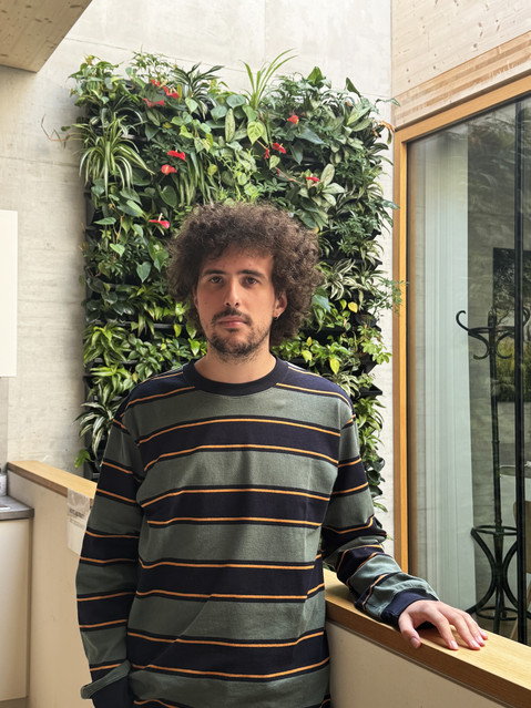 Porträt von Lorenzo Maccarone in einem grün-schwarz gestreiften Langarmshirt, der sich an ein Geländer vor einer vertikalen, begrünten Pflanzenwand im Institutsgebäude lehnt.