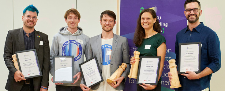 Foto der Gewinner*innen 2025 mit Urkunden und Holzawards in Form von Leuchttürmen. Von links nach rechts: Thomas Filek, Max Nutz, Nicolas Roux, Friederike Frieß, Max Pramreiter.