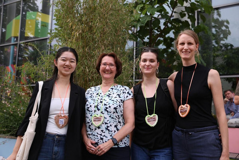 The photo shows a group of four smiling women standing next to each other outdoors, in front of a modern building with large glass windows and surrounded by plants (including bamboo). From left to right, they are: Liting Yuan, Astrid Gühnemann, Georgia Charalampidou, and Corinna Mölkner. As a special shared detail, all four women are wearing decorated gingerbread hearts on ribbons around their necks.