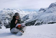 Gebirgslandschaft im Winter, im Vordergrund Studentin im Schnee sitzend und ein Buch lesend