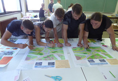 Eine Gruppe von fünf Studierenden beugt sich gemeinsam über mehrere große Tische. Sie arbeiten intensiv an farbigen Lageplänen und Postern, die verschiedene Varianten von Kreisverkehren und Kreuzungen zeigen.