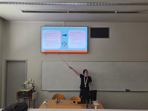 Olivia Gold standing in a seminar room, using a long pointer stick to indicate a BOKU presentation slide titled "A low-carbon mobility transition – Who gains? Who loses?" on a wall-mounted display.   