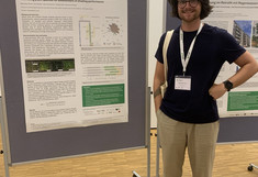 Max Poiss in a black T-shirt, standing sideways in front of his poster