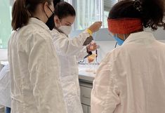 Teilnehmerinnen in weißen Labormänteln vermischen die Probe aus Obst mit einem Glasstab