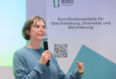 Preisträgerin Melanie Pichler mit einem Mikrophon vor dem Rollup der Koordinationsstelle beim vorstellen ihrer Lehrveranstaltung