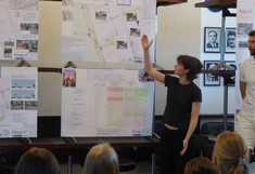 Eine Studentin steht mit erhobenem Arm vor dem Poster „Evaluation“ der Gruppe 2. Sie erklärt dem Publikum die systematische Bewertung der Planungsstufen anhand einer farbcodierten Kriterien-Matrix.