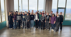 Group photo of the Institute for Transport Studies (BOKU) team standing in a sunlit seminar room with a scenic view of a town and mountains in the background. A group of approximately 25 people is standing together in two rows in a spacious, bright room with a wooden floor. The group is dressed in casual to smart-casual attire. In the background, large floor-to-ceiling windows offer an expansive view of a valley, a town, and distant mountains under a light sky. The room features teal-colored wall accents and neutral-toned curtains.