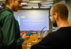 Zwei Gäste im Gespräch, unscharf, im Fokus ist die Leinwand, auf der "BOKU Leading Lights, Preis für Wissenschaftskommunikation von Nachwuchsforscher*innen" zu lesen ist