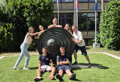 Gruppenfoto der international gemischten „Gruppe 1“: Fünf Studierende und ihr Betreuer Wolfgang J. Berger posieren gut gelaunt auf einer Rasenfläche um und auf einer großen, kreisförmigen, dunklen Metallskulptur.