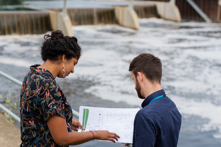 Eine Frau und ein Mann blicken auf einen Plan. Im Hintergrund ist ein Querbauwerk in einem breiten Fluss zu erkennen.