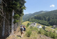 The students walk steeply downhill along the edge of the forest; below, an old church with a cemetery and a few houses can be seen.