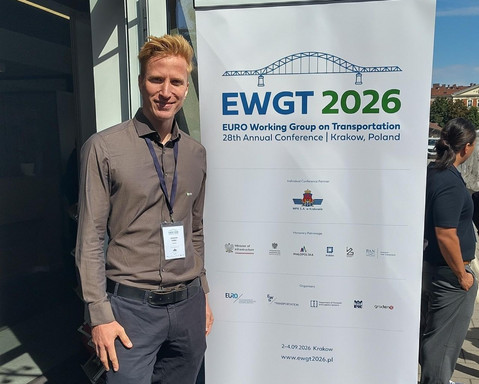 Johannes Müller steht mit Konferenz-Badge lächelnd neben einem Roll-up-Banner der Verkehrsforschungskonferenz EWGT 2026 in Krakau im Außenbereich.