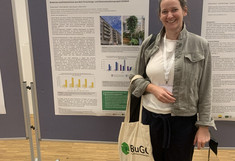 Laughing Ulrike Pitha stands in a grey-green jacket and black trousers in front of her poster