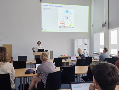 Prof. Astrid Gühnemann hält einen Vortrag an einem Rednerpult in einem Seminarraum vor Publikum, während auf einer großen Leinwand eine BOKU-Präsentationsfolie mit einem Systemdiagramm zum Thema Feedback-Schleifen projiziert wird.