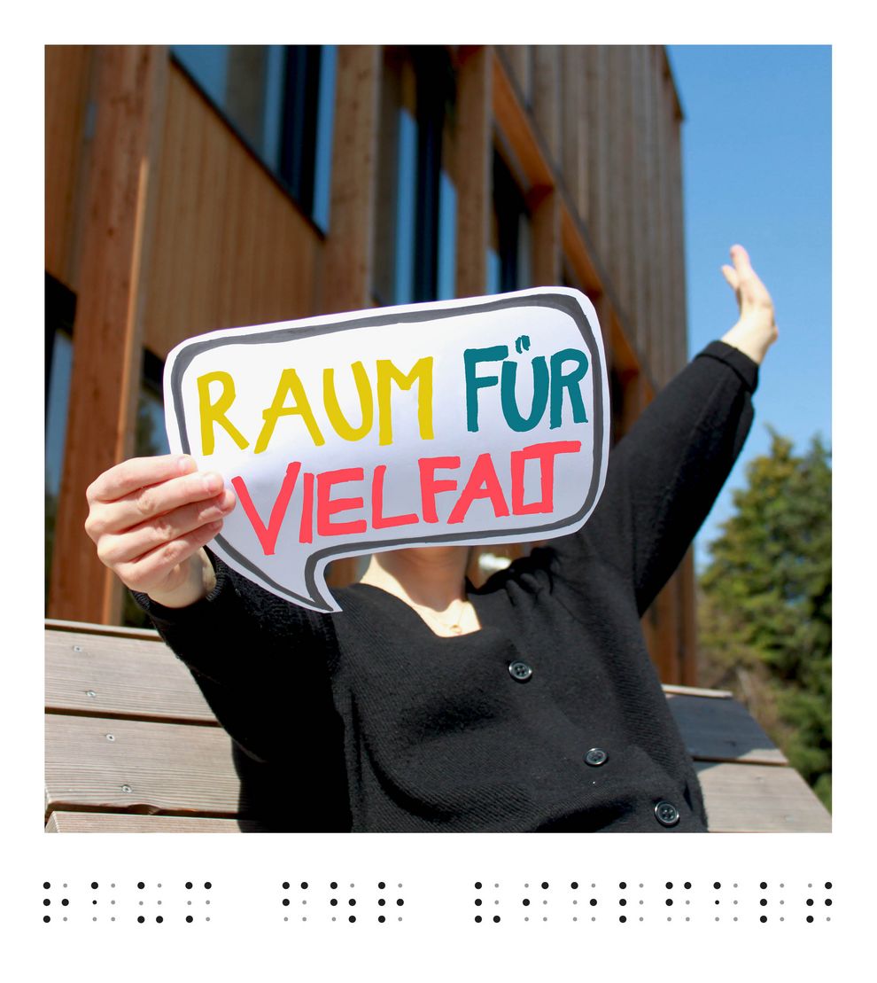 Foto mit Person, welche eine Sprechblase mit der Aufschrift "Raum für Vielfallt" hält. Im Hintergrund das Ilse-Wallentin-Haus . 