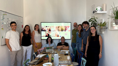 9 Personen in einem Besprechungsraum stehen um einen Tisch und lächeln in die Kamera. Dahinter ein Monitor mit Videokonferenzteilnehmer *innen. 