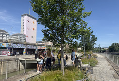 The group is standing in the shade of a tree and on a lock wall with planted areas.