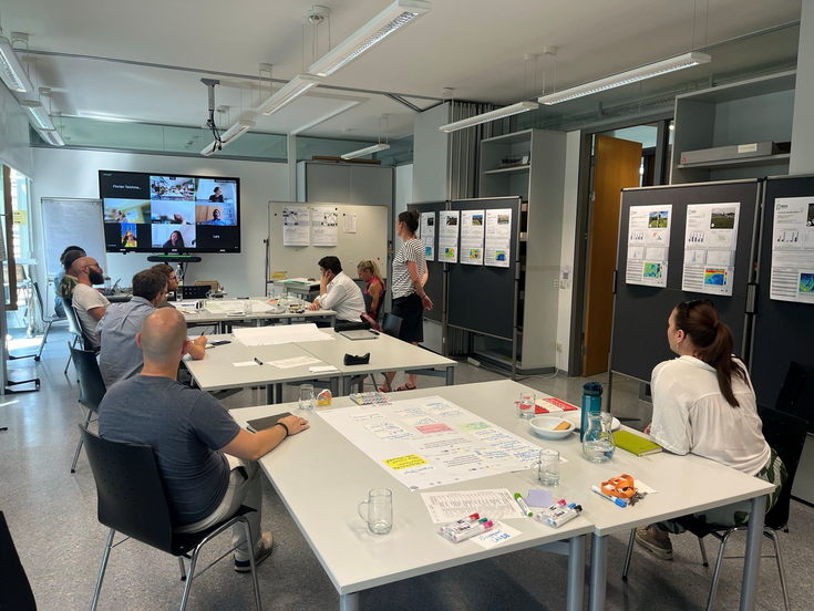 Seminarraum mit einigen Teilnehmer*innen. Plakate an den Wänden, beschriftete Flipcharts auf den Tischen und Videoteilnehmer*innen am großen Monitor im Hintergrund.
