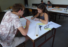 Zwei Studierende sitzen an einem Tisch und arbeiten konzentriert an einem großformatigen Papierplan. Ein Student hält einen Stift, die Studentin zeichnet mit einem Lineal und einem Kurvenlineal auf dem Papier.