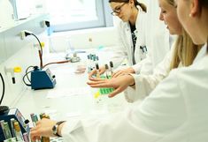 Das Foto zeigt mehrere Frauen in weißen Labormäntel vor einem Labortischaufbau und Laborgeräten