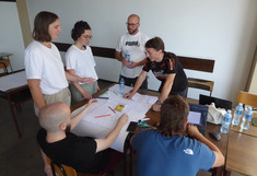 Eine internationale Gruppe von Studierenden steht um einen Tisch versammelt und diskutiert gemeinsam an einem Projektplan. Ein Teilnehmer arbeitet am Laptop, während die anderen den Entwurf begutachten.