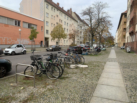 Straße mit Parkenden Autos, breiter Gehweg mit parkenden Fahrrädern, Wohn und Fabriksgebäude.