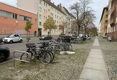 Street with parked cars, wide sidewalk with parked bicycles, residential buildings and a small factory